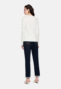 White long-sleeve top with rounded neckline, paired with dark blue jeans featuring decorative stitching on back pockets, and light-colored heels.