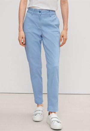 Pantalones ajustados de color azul claro, fabricados en una mezcla de algodón. Presentan bolsillos delanteros y un cierre de un solo botón. Se llevan con zapatillas blancas.