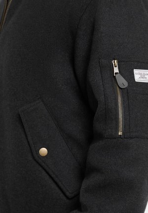 Giacca nera in misto lana con tasca con zip, dettagli dorati e tasca laterale a pattina. Il tessuto ha un aspetto liscio e strutturato.