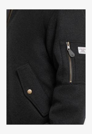 Melnas vilnas maisījuma jaka ar rāvējslēdzēja kabatu, zelta akcentiem un sānu kabatu ar flapību. Audumam ir gluda, teksturēta virsma.