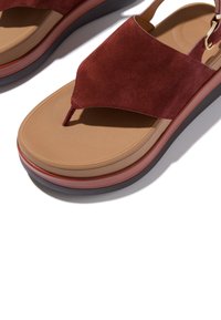 Close-up van een sandaal met een bruine leren zool, een donkerrode suède brede band en teenbandje, en een verstelbare enkelband met gouden gesp.