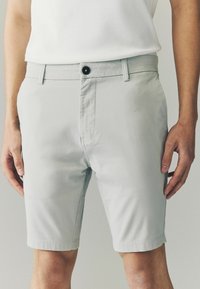 Lys grå shorts lavet af glat materiale. Har en frontknaplukning, bæltestropper og en lige pasform, der slutter lige over knæet.