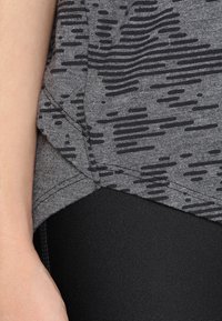 Grå idrottstopp med ett texturerat, abstrakt mönster i svart. Har en böjd nederkant och är kombinerad med svarta leggings.