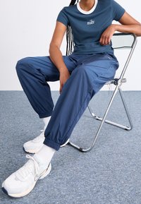 Osoba sedící na kovové židli, oblečená v modrém tričku Nike, volných modrých kalhotách, bílých ponožkách a bílých teniskách Nike na šedém koberci.