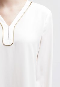 Hvid bluse med lange ærmer, V-udskæring kantet med guld. Blød stofstruktur, der har en subtil glans og rene sømdetaljer.
