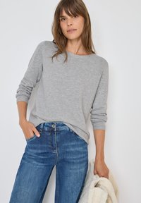 Maglietta grigia chiaro a maniche lunghe con scollatura rotonda, abbinata a jeans blu a gamba dritta. Il modello tiene un giacchino texturizzato color crema.