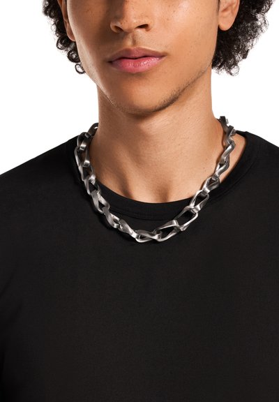 Collier en chaîne métallique argentée avec des maillons torsadés, porté sur un t-shirt noir. Gros plan sur la zone du cou, mettant en valeur la texture lisse et le design.