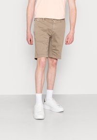 Pantaloni corti in cotone beige con vestibilità dritta, dotati di zip e tasche frontali. Abbinati a sneakers bianche e calze alla caviglia.