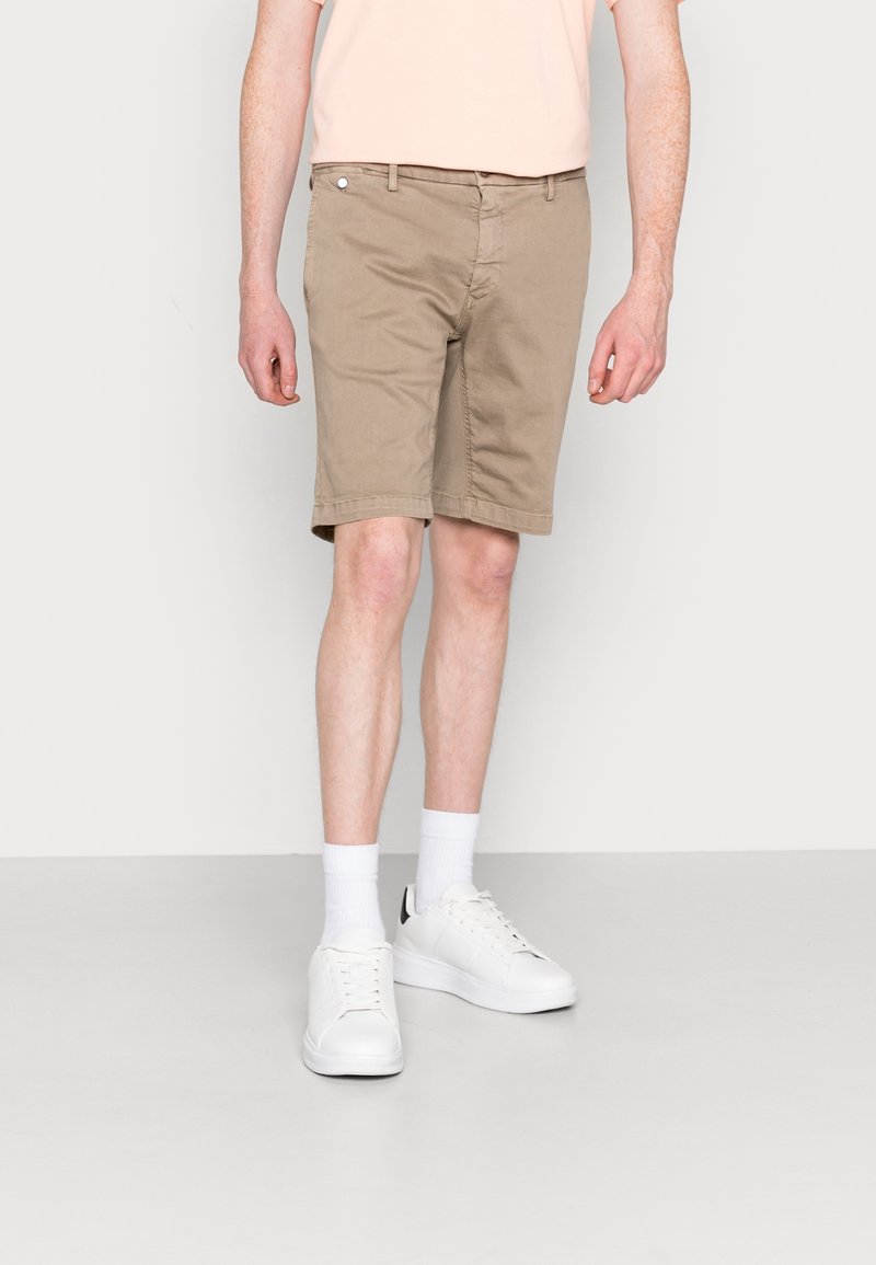 Pantaloni corti in cotone beige con vestibilità dritta, dotati di zip e tasche frontali. Abbinati a sneakers bianche e calze alla caviglia.
