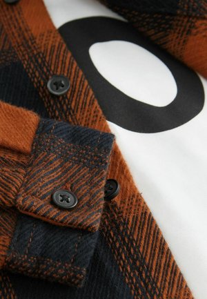 Chemise en flanelle orange et marine avec un motif à carreaux, dotée de boutons noirs et d'un t-shirt blanc en dessous affichant un graphique noir audacieux.