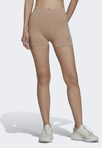 Shorts de sport beiges avec une taille haute et une texture lisse. Présente un petit logo argenté sur la jambe gauche. Portés avec des chaussures de sport blanches.