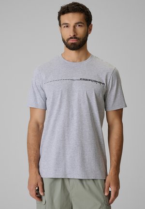 Mężczyzna z krótkimi ciemnymi włosami i brodą, ubrany w jasnoszary t-shirt z czarnym tekstem "4F Sportswear" i jasnoliwymi spodniami na jednolitym tle.