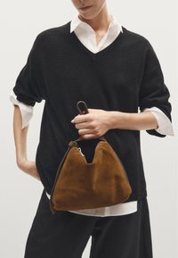 Brown suede bag with a triangular shape and a top handle, held by a person wearing a black sweater over a white collared shirt.
