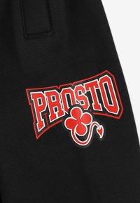 Czarna tkanina z wyraźnym czerwono-białym logo "PROSTO". Zawiera grafikę czerwonego trójlistnego koniczka oraz design ogonem diabła.