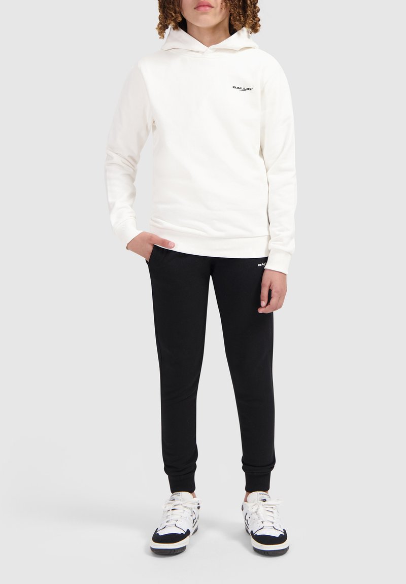 Witte sweatshirt met een capuchon en zwart logo, gecombineerd met zwarte joggingsbroeken. De outfit heeft een relaxte pasvorm en is gemaakt van zachte stof.