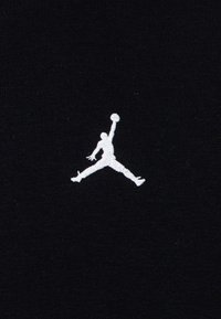 La tela negra presenta un logo bordado en blanco de una figura humana en una posición de salto, sosteniendo un balón de baloncesto por encima de su cabeza.