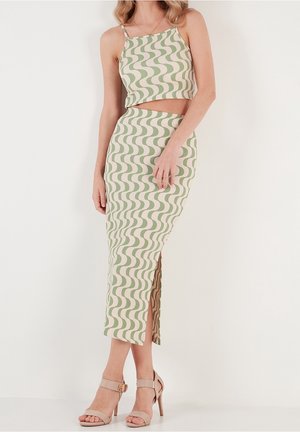 Kvinde iført en grøn og cremefarvet crop top med bølgemønster og matchende højhøj taljeskørt med sideslid samt beige højhælede sandaler.