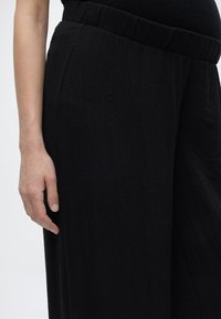 Το αριστερό χέρι ακουμπά δίπλα σε μαύρο, υφασμάτινο, φαρδύ παντελόνι με ελαστική μέση, πάνω σε απλό, ανοιχτόχρωμο φόντο.