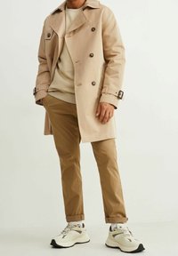 Beige dvoubarevný trenčkot s tmavými knoflíky, béžový svetr pod ním, světle hnědé kalhoty a bílé tenisky s černými akcenty.