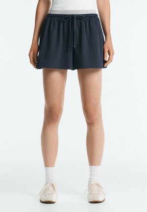 Femme portant un short à rayures bleu marine avec cordon de serrage, des chaussettes blanches et des baskets blanches, debout devant un fond clair uni.