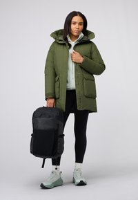 Giubbotto puffer verde oliva con cappuccio, tasche frontali, indossato sopra un leggero pile menta. Zaino nero tenuto in una mano, scarpe leggere.