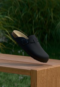 Μαύρα παπούτσια τύπου clog με λουρί και αγκράφα, με λείο συνθετικό επάνω μέρος και υφή, καλουπωμένη σόλα, τοποθετημένα πάνω σε ξύλινη επιφάνεια.