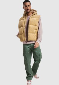 Chaleco de pana acolchado color marrón claro con capucha, llevado sobre un sudadera rosa claro. Combinado con pantalones cargo verdes y zapatillas de color claro.