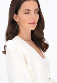 Biały sweter z prążkowanej tkaniny z falbanką na ramieniu, z głębokim dekoltem w kształcie litery V oraz dużym zapięciem na guzik.