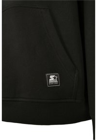Poche de sweat à capuche noire avec écusson du logo Starter Black Label cousu près de l'ourlet côtelé sur un tissu foncé.
