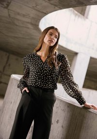 Blusa nera a pois realizzata in tessuto leggero e trasparente con maniche lunghe a sbuffo, abbinata a pantaloni neri a vita alta.