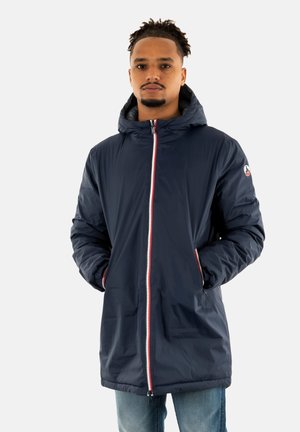 Cappotto impermeabile blu navy con cappuccio, dotato di chiusura frontale con zip, accenti rossi e polsini elastici. Texture liscia e design lungo fino al ginocchio.