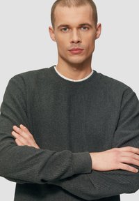 Jeune homme avec des cheveux clairs courts et des yeux bleus portant un pull en tricot gris foncé, se tenant les bras croisés devant un fond uni.