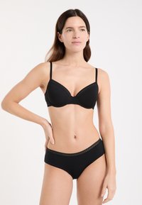Donna che indossa un set coordinato di reggiseno e mutandine nere, in piedi con una mano sui fianchi, che guarda leggermente di lato contro uno sfondo semplice.