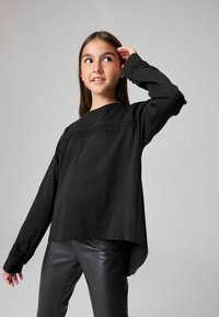 Must läng sleeves särk pehmest materjalist, ümarate lõhenemistega kaelusega ja dekoratiivse tekstuuriga riba rinnal, kantud mustade pükstega.
