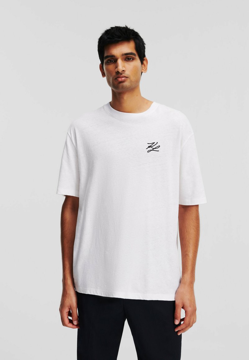 T-shirt bianca in cotone con vestibilità rilassata, scollatura rotonda e maniche corte. Presenta un logo ricamato nero sul lato sinistro del petto.