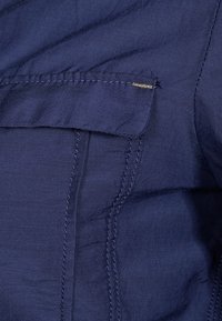 Chemise bleu marine avec une poche poitrine, texture lisse, couture visible et un petit accent doré près du bord de la poche.