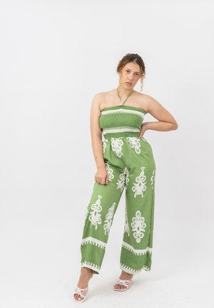 Frau trägt einen grünen trägerlosen Jumpsuit mit weißen Blumenmustern und weißen Sandaletten mit Absatz, steht vor einem einfarbigen hellen Hintergrund.