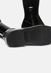 Botas negras brillantes hasta el tobillo con una cremallera en el lateral y una suela de goma texturizada. El tacón es plano para mayor estabilidad.