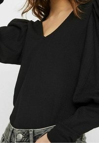 Black, textured long-sleeve top with a V-neckline, featuring puffed shoulders. Paired with gray high-waisted jeans.