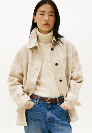 Beige geruite jas met knopen, gelaagd over een crèmekleurige kabeltrui. Blauwe hoogzittende jeans met een bruine leren riem.