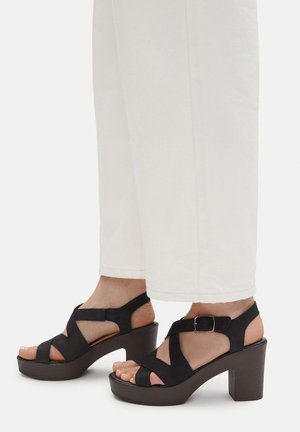 Schwarze Plateausandalen aus Wildleder mit gekreuzten Riemen und einem Knöchelriemen, ausgestattet mit einer klobigen Sohle und offenen Zehen, kombiniert mit cremefarbenen Hosen.