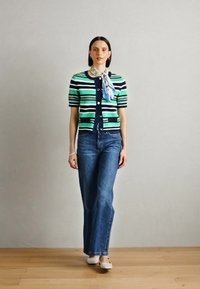 Cardigan à rayures vertes et bleu marine avec des manches courtes, fermeture à boutons, associé à un jean bleu évasé et une écharpe bleu clair nouée au cou.