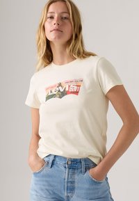 T-shirt en coton couleur crème avec un imprimé logo Levi's rétro multicolore sur le devant. Il a des manches courtes et un col rond.