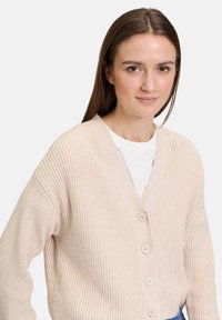 Beige gestrickte Strickjacke mit V-Ausschnitt, fünf Knöpfen und geripptem Material. Über einem weißen T-Shirt getragen, zeigt sie lockere Ärmel und ein lässiges Design.