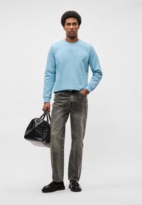 Sweatshirt de algodão azul claro com gola redonda, calças de ganga cinzentas e sapatos de couro pretos. A segurar uma mala de viagem preta texturizada.