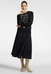 Donna che indossa un maglione nero ricamato con fiori, una gonna nera a trapezio, stivali con tacco beige e orecchini a forma di stella, posando con la mano vicino al viso.