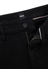 Pantalones de mezclilla negra con una etiqueta BOSS visible, un cierre de cremallera plateado, un cierre de botón y costuras en contraste a lo largo de la cintura.