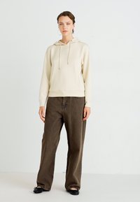Kreemiga kapuutsiga sweatshirthoodie, millel on nöörid ja soonikäär, koos pruunide laia jala pükstega. Mustad lamedad kingad ja minimaalsed aksessuaarid täiendavad komplekti.