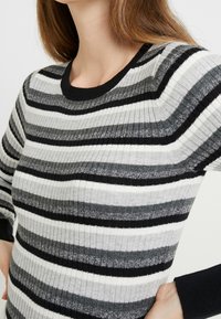 Sweter z dzianiny prążkowanej z poziomymi paskami w odcieniach czerni, szarości i bieli, z czarnym okrągłym dekoltem oraz długimi rękawami.