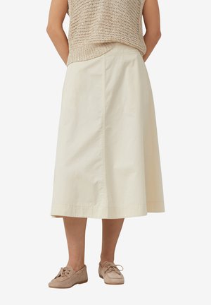 Femme portant une jupe crème longueur cheville avec un haut en maille beige et des chaussures taupe à lacets, debout les mains derrière le dos.